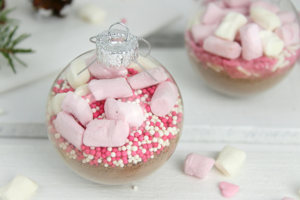 Trinkschokolade mit bunten Streuseln, Zuckerstange und Marshmallows in einer Christbaumkugel