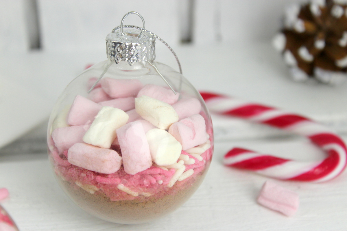 Trinkschokolade mit bunten Streuseln, Zuckerstange und Marshmallows in einer Christbaumkugel